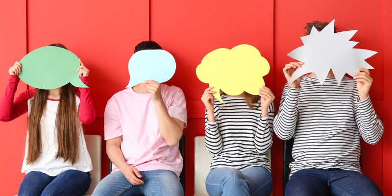 Četri cilvēki sēž pie sarkanas sienas, aizklājot savas sejas ar dažādu krāsu runas un domu burbuļiem – zaļu, gaiši zilu, dzeltenu un baltu.