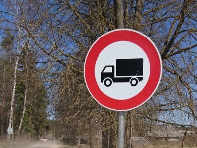 Apaļa ceļa zīme ar sarkanu apmali un melnu kravas automašīnas siluetu baltā fonā, kas norāda kravas automašīnu satiksmes aizliegumu. Fonā kaili koki, zilas debesis un grants ceļš.