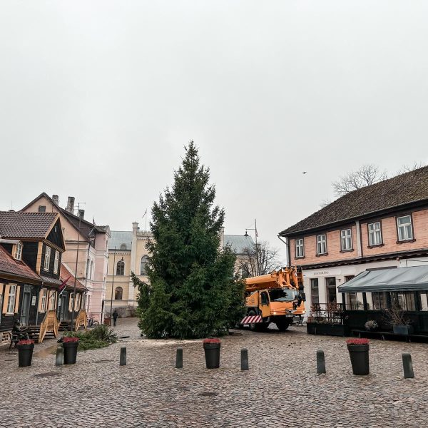 Liela Ziemassvētku egle Kuldīgas Rātslaukumā, tai blakus oranža celtņa mašīna, ko ieskauj vēsturiskas ēkas. Uz vienas no tām redzams uzraksts "RESTORĀNS".
