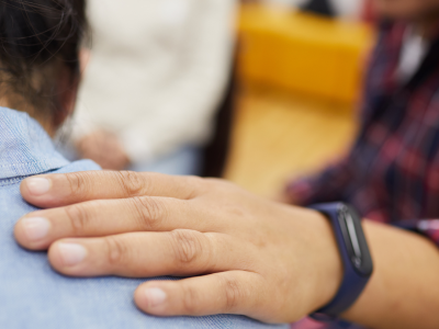 Roka uz cilvēka pleca, sniedzot atbalstu. Uz rokas ir viedpulkstenis, fonā neskaidri redzamas citas personas.