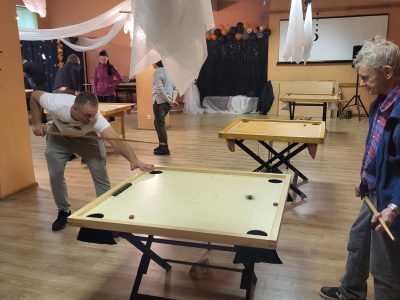 Vīrietis baltā T-kreklā spēlē novusu, tēmējot ar kiju pa melnu ripu uz gaiša galda. Viņam blakus labajā pusē stāv vecāks vīrietis zilā jakā ar rūtainu kreklu, turot kiju un vērojot spēli.