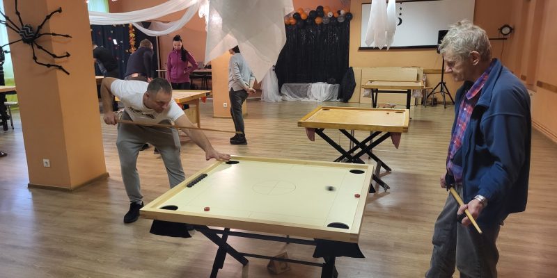 Vīrietis baltā T-kreklā spēlē novusu, tēmējot ar kiju pa melnu ripu uz gaiša galda. Viņam blakus labajā pusē stāv vecāks vīrietis zilā jakā ar rūtainu kreklu, turot kiju un vērojot spēli.