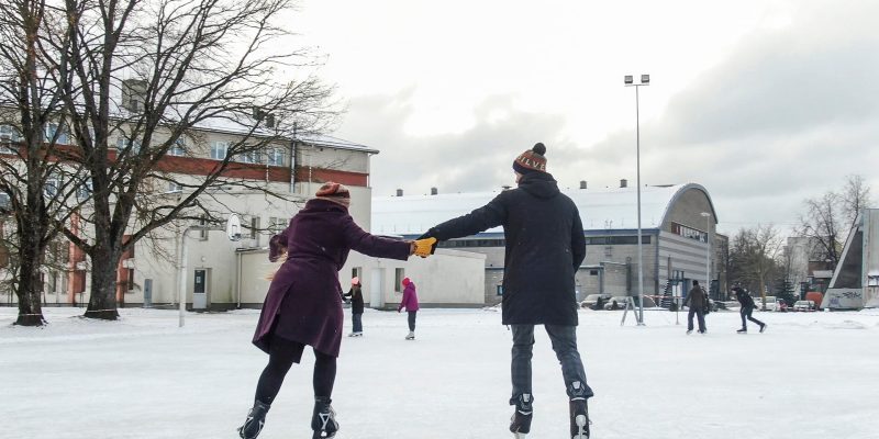 Pāris, turoties pie rokām, slido āra slidotavā ziemas dienā. Vīrietis valkā cepuri ar uzrakstu "LV", bet sieviete cepuri ar uzrakstu "SILVE". Fonā redzamas ēkas un citi slidotāji. Kuldīgas slidotava.