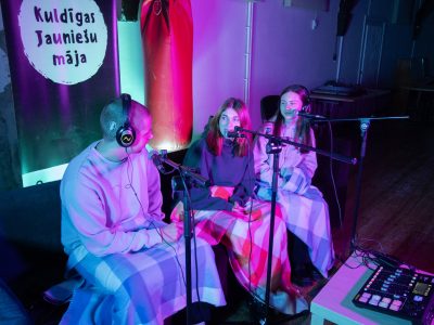 Trīs jaunieši, divas sievietes un vīrietis, sēž zem segām, runā mikrofonos un valkā austiņas, ierakstot podkāstu purpursarkanās un zilās gaismās. Fonā redzams plakāts ar uzrakstu "Kuldīgas Jauniešu māja".