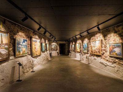 Plaša gleznu izstāde šaurā, garenā telpā ar neapstrādātām akmens sienām un tumšu griestiem, kur gleznas izgaismo prožektori.