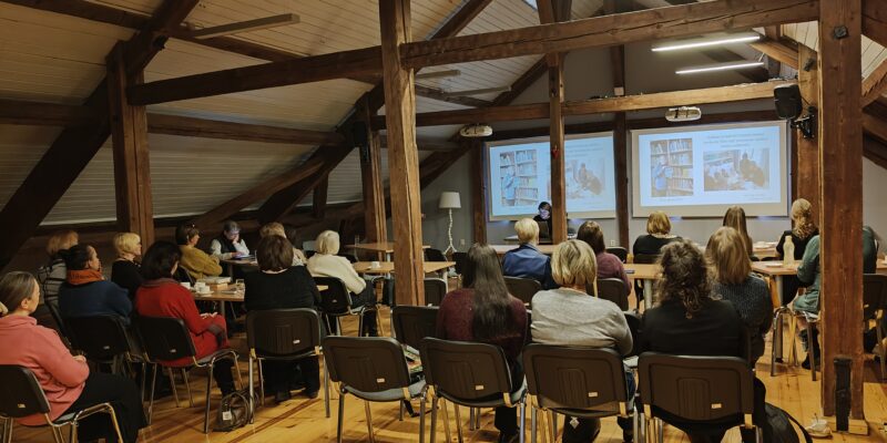 Bēniņu telpa ar atsegtām koka sijām, kurā pieaugušo grupa sēž krēslos, vērojot prezentāciju. Priekšā redzami divi projektoru ekrāni, kuros attēloti grāmatu plaukti un cilvēki, kas lasa vai strādā pie galdiem.