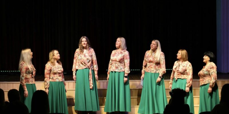 Septiņas sievietes saskaņotos tērpos – ziedu raksta blūzēs un garos, zaļos svārkos – uzstājas uz skatuves. Viņas stāv rindā pret tumšu fonu, dažas skatās viena uz otru, dažas uz priekšu, un viena sieviete dzied ar atvērtu muti. Priekšplānā redzami skatītāju silueti.