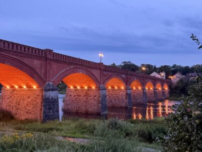 Vecs sarkano ķieģeļu velvju tilts ar akmens balstiem, kas krēslā izgaismots ar silti oranžām gaismām, atspoguļojas upē. Priekšplānā zāļaini krasti.