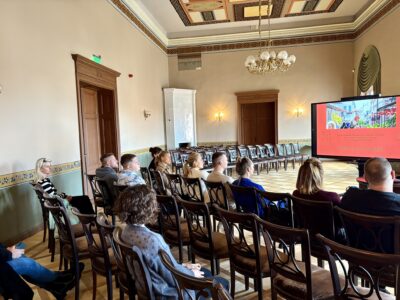 Grupa cilvēku sēž greznā zālē un skatās uz lielu ekrānu, kurā redzams ielas attēls