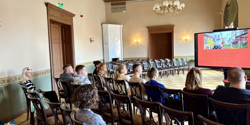 Grupa cilvēku sēž greznā zālē un skatās uz lielu ekrānu, kurā redzams ielas attēls