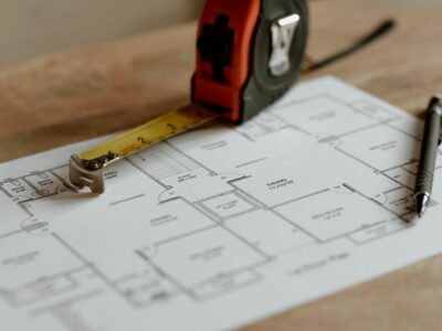 Mājas plāns uz papīra ar izvilktu mērlenti un pildspalvu. Plānā redzami telpu apzīmējum